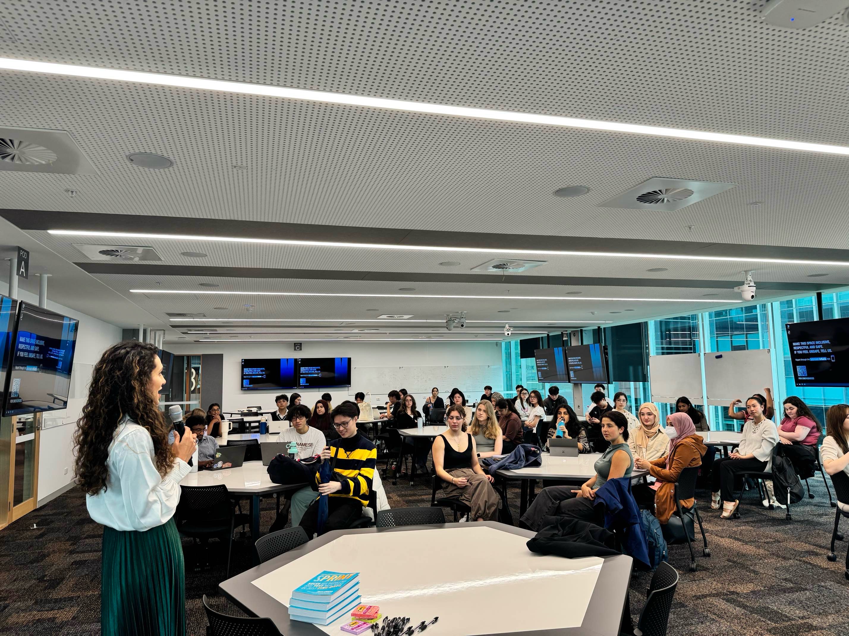 A speaker addresses a large group of university students seated in a modern classroom, with screens and whiteboards in the background.
